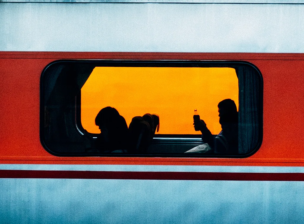 Silhouettes of passengers seen through a train window against a bright orange sky, framed by a red carriage.