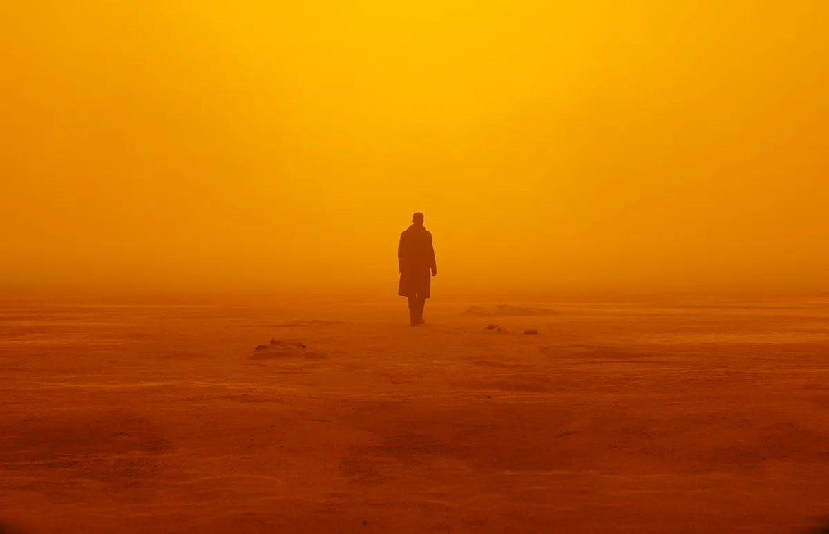 A lone figure in a long coat stands in thick orange haze on a bare, dusty plain.
