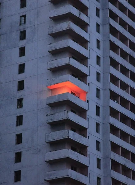 A grey concrete tower block with stacked balconies, one balcony glowing orange-red.